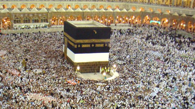 A large crowd of people around a black square structure with Kaaba in the background  Description automatically generated with low confidence
