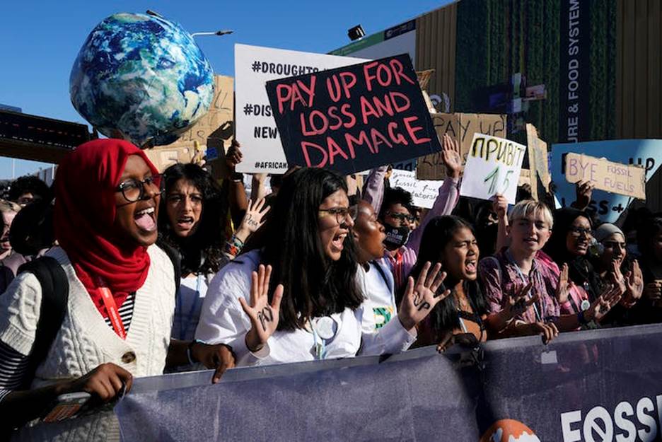 Young people from many countries shout and wave signs reading 'pay up for loss and damage' at a small outdoor protest.