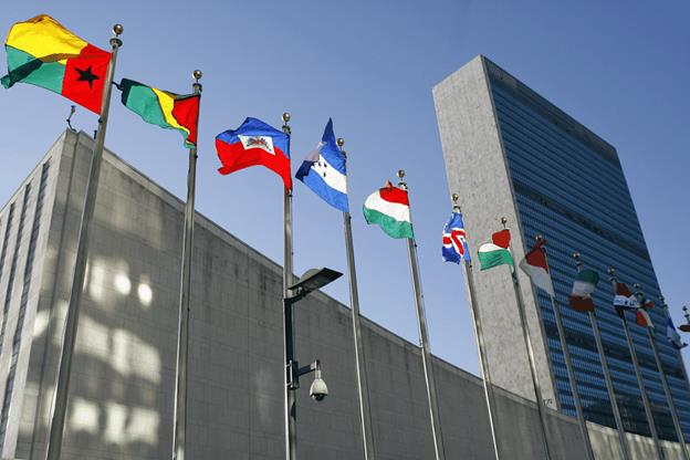 A group of flags on poles with Headquarters of the United Nations in the background  Description automatically generated