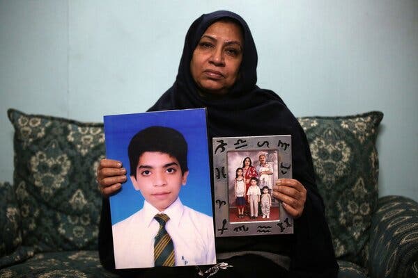 A woman in black garb sits on a couch while holding up two photographs: an image of a child and a family portrait.