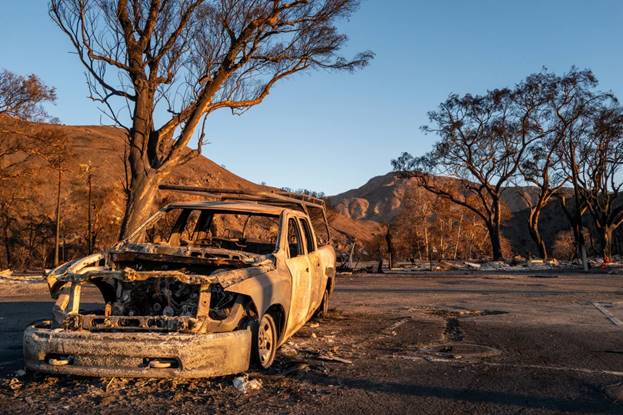 With paint charred and grill empty and showing the shell of the exposed engine, a vehicle sits on what appears to be an empty parking lot surrounded by burnt trees.