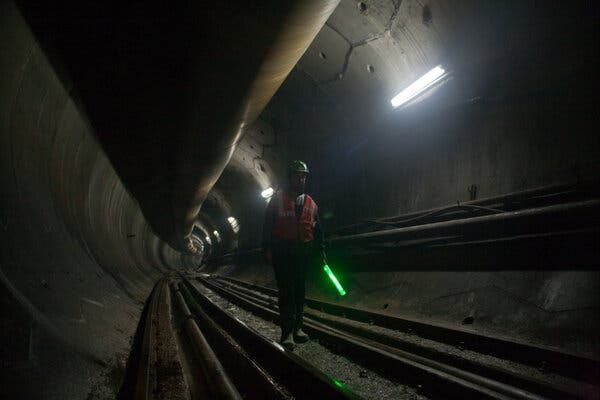 A worker in a hard hat and high-vis vest, holding a light stick, walks through a dimly lit tunnel.