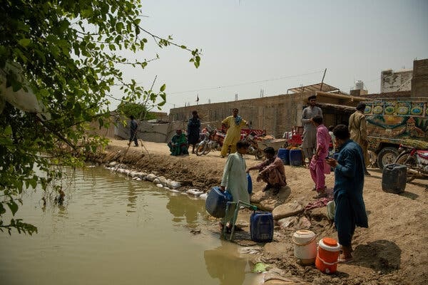 People stand on sandy banks next to a stream, many with plastic containers to hold water.